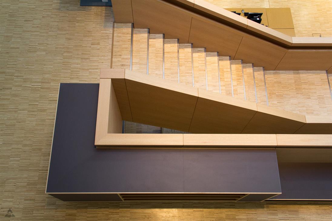 Blick senkrecht herunter aus dem Obergeschoss der Bibliothek am Campus Derendorf. Die Holztreppe in das Untergeschoss ist bildfüllend abgebildet. Stufen und Geländer bilden geometrische Formen.