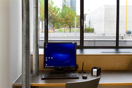 A computer workplace on a brown surface. Next to the workplace, metal tubes range from the floor to the ceiling. A window behind the workplace shows a view to autumn trees.