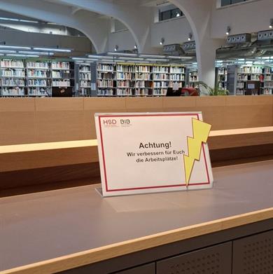 Foto: Schild auf dem Medienring "Achtung! Wir verbessern für Euch die Arbeitsplätze." mit einem gelben Blitz. Unterhalb Schließfächer, im Hintergrund Buchregeale und Deckengewölbe.