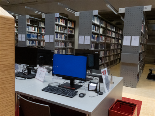 Foto: Im Vordergrund Tische mit mehreren PC-Arbeitsplätzen inkl. Monitoren und Tastaturen, im Hintergrund Buchregale.
