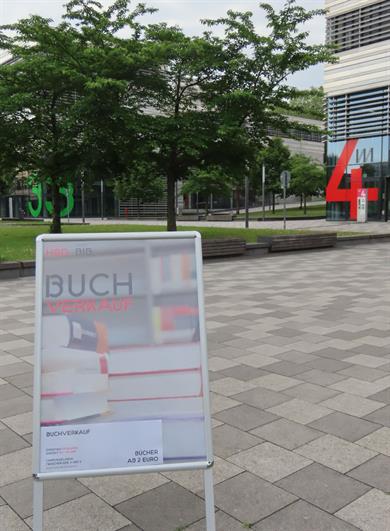 Foto: im Vordergrund Plakataufsteller zum Buchverkauf, im Hintergrund Grünanlage und Gebäude 3+4 der HSD