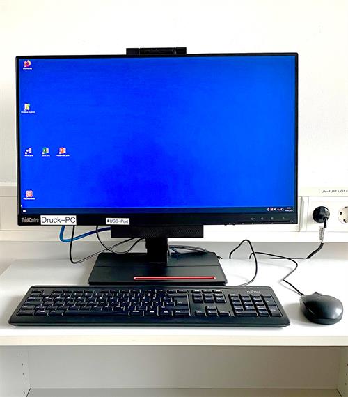 Foto: PC mit Monitor und Maus steht auf einem weißen Tisch. Auf dem Bildschirm ist der blaue Hintergrund mit sechs Icons zu sehen. Beschriftet ist der Monitor mit Druck-PC.