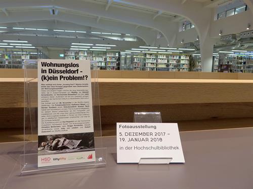brochures of the photo exhibition Housing First in the University Library are placed on the handrail. In the background one can see the reading room and bookshelves.