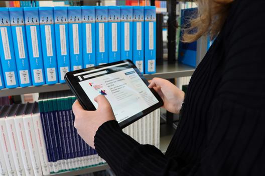 Foto: Nahaufnahme einer Person von hinten, die ein Tablet hält und darauf zu lesen scheint. Im Hintergrund ist in der Nahaufnahme ein Buchregal.