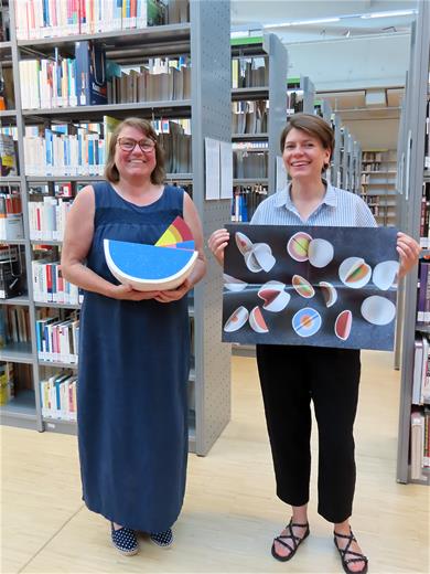 Foto: Vor Buchregalen stehen zwei Frauen, die ein Poster und mehrere wie Melonenstücke geformte Pappkartons halten.