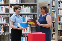 Foto: Zwei Frauen stehen vor Buchregalen und schauen sich mehrere wie Melonenstücke geformte Pappboxen an.