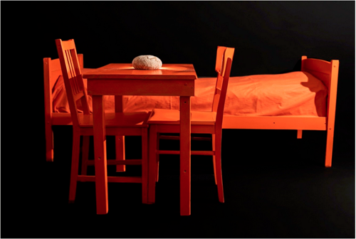 Foto: vor schwarzen Hintergrund steht ein oranges Bett und ein oranger Tisch mit zwei Stühlen. Auf dem Tisch liegt ein rundes Brot.