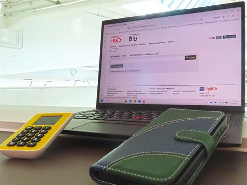 Foto: Auf der Tastatur eines Laptops liegt links ein Authentificator-Gerät und rechts ein Handy mit einer grünen Hülle. Auf dem Bildschirm ist die Einstiegsseite des Katalog Plus der BIB zu sehen.