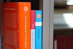 Foto: Nahaufnahme eines Buchregals mit drei farbigen Büchern mit Titeln rund um DIN-Normen. Hintergrund verschwommen.